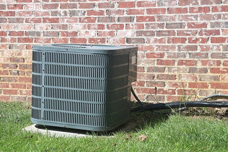 Blog Title: Top Causes of AC Failure Photo: Home Air Conditioner Condenser coil sitting in front of brick wall